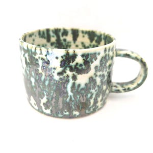 May include: A ceramic mug with a distinctive speckled glaze in shades of green, teal, and black. The mug features a rounded body, a robust handle, and a slightly irregular rim. The interior is a creamy white colour.