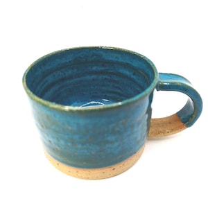 May include: Handmade ceramic mug with a teal blue glaze and a natural tan base and handle. The mug has a textured surface and a curved handle. Ideal for tea, coffee, or any warm drink.