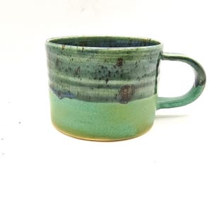 May include: Handmade ceramic mug with a rounded shape and a large handle. The mug features a two-tone glaze, with a light green base and a darker, mottled green upper section. The mug is suitable for coffee, tea, or other beverages.