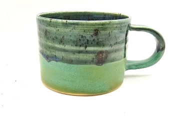 May include: Handmade ceramic mug with a rounded shape and a large handle. The mug features a two-tone glaze, with a light green base and a darker, mottled green upper section. The mug is suitable for coffee, tea, or other beverages.