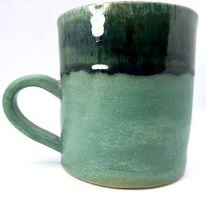 May include: A green ceramic mug with a dark green and blue glaze. The mug has a rounded handle and a wide base.
