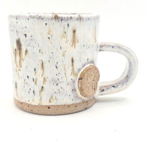 May include: A ceramic mug with a speckled white and tan glaze featuring brown vertical streaks. The base is unglazed, showing a textured tan clay. A circular clay medallion with an embossed spiral design is attached where the handle meets the body.