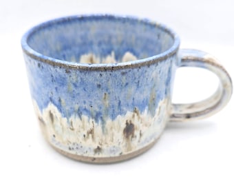 May include: A handmade ceramic mug with a blue and white glaze. The mug has a rounded shape with a handle and a capacity of approximately 12 ounces. The glaze has a textured appearance, with variations in color and tone.