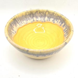 May include: A handmade ceramic bowl with a bright yellow interior and a cream-coloured exterior. The rim features a textured, grey and cream-coloured glaze. The bowl is round and has a glossy finish.