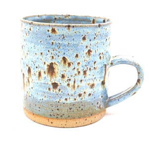May include: A handcrafted ceramic mug with a speckled blue glaze and brown flecks. The mug features a rounded form, a robust handle, and an unglazed base. Ideal for tea or coffee, this unique mug adds a touch of rustic charm.