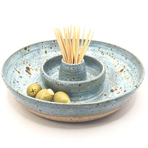 May include: A handcrafted ceramic serving platter with a pale blue glaze and brown flecks. It features a central section for toothpicks and a surrounding area for olives. The rim is a natural, unglazed colour.