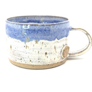 May include: Handmade ceramic mug with a blue and white speckled design. The mug has a rounded handle and a small, textured emblem on the side. The base is a natural, unglazed colour.
