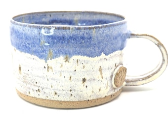 Op de afbeelding: Handgemaakte keramische mok met een blauw-wit gespikkeld ontwerp. De mok heeft een afgerond handvat en een klein, getextureerd embleem aan de zijkant. De basis is een natuurlijke, ongeglazuurde kleur.