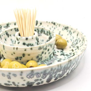May include: A speckled ceramic olive serving dish with a central toothpick holder. The dish is off-white with dark grey and teal speckles. Green olives are in the dish. A bundle of wooden toothpicks stands in the centre.