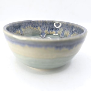 May include: A handmade ceramic bowl with a unique glaze. The bowl has a light blue base, transitioning to a creamy white middle, and a dark blue rim with gold flecks. The bowl has a slightly irregular shape.
