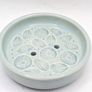 May include: A light blue ceramic soap dish with a floral design. The dish has three drainage holes.