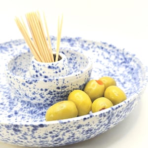 May include: A white ceramic serving dish with a blue speckled pattern, featuring a central raised bowl for toothpicks and a surrounding area for snacks. A cluster of green olives is placed on the dish, and a bundle of wooden toothpicks is inside the centre bowl.