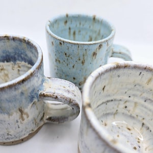 May include: Three handmade ceramic mugs in varying shades of blue and white. The mugs have a speckled glaze and visible brushstrokes, with rounded handles. The mugs are of different sizes and shapes, showcasing a rustic, artisanal aesthetic.