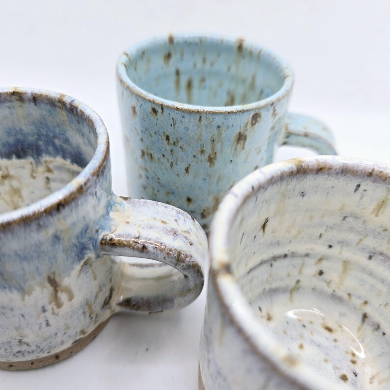 May include: Three handmade ceramic mugs in varying shades of blue and white. The mugs have a speckled glaze and visible brushstrokes, with rounded handles. The mugs are of different sizes and shapes, showcasing a rustic, artisanal aesthetic.