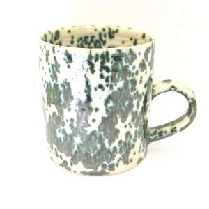 May include: A ceramic mug with a speckled glaze in white, teal, and dark green. The mug has a cylindrical shape and a rounded handle. The speckled pattern covers the entire surface of the mug, giving it a unique appearance.
