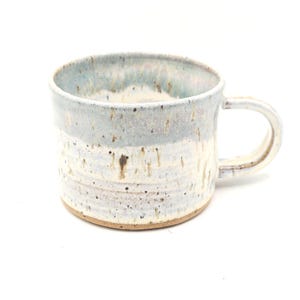 May include: A ceramic mug with a light blue and white glaze, brown speckles, and drips. The mug has a rounded body and a handle. Suitable for hot drinks, the mug is handmade and has a rustic appearance.
