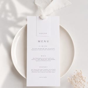 Op de afbeelding: Een witte rechthoekige menukaart met zwarte tekst met de naam Louise, secties voor voorgerecht, hoofdgerecht en dessert, en een wit gerafeld lint aan de bovenkant, rustend op een effen wit bord. De tekst bevat een lijst met gerechten.