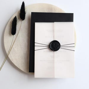 May include: A white invitation with a black wax seal and black string. The invitation is on a white marble surface.