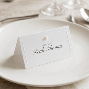 Może przedstawiać: Biała wizytówka z imieniem "Leah Thomas" elegancką czcionką, zwieńczona perłą. Karta znajduje się na białym talerzu, z sztućcami i szkłem w tle. Nad imieniem widnieje słowo "WELCOME".