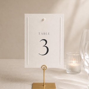 Può includere: Un segnatavolo rettangolare bianco con la scritta TABLE 3 in carattere serif nero. La carta presenta linee verticali in rilievo sui lati, un piccolo dettaglio a perla nella parte superiore centrale ed è sostenuta da un supporto in metallo oro.