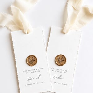 Könnte beinhalten: Zwei rechteckige weiße Papieranhänger mit Büttenrand, jeweils mit einem goldenen Wachssiegel mit Blumenmotiv versehen. Der Text lautet: This seat is kindly reserved for Daniel, Mother of the Bride, und Helen, Mother of the Bride. Cremefarbene Bänder sind befestigt.