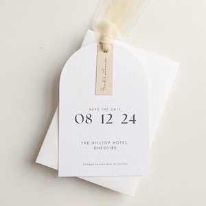 May include: A white Save the Date card with an arched top, featuring the text "08 12 24" and "The Hilltop Hotel, Cheshire". A small tag with "Red & Louise" is attached with a cream ribbon. A white envelope is behind the card.