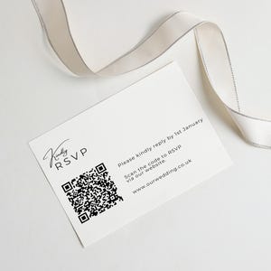 Puede incluir: Una tarjeta RSVP rectangular blanca con texto negro que dice Kindly RSVP, Please kindly reply by 1st January, Scan the code to RSVP via our website y www.ourwedding.co.uk, junto a un c&oacute;digo QR negro y una cinta con borde plateado.