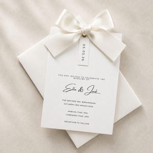 Puede incluir: Una tarjeta de invitaci&oacute;n de boda blanca con texto negro que dice Ella & Jack, The Oaktree Inn, Birmingham, Saturday 3rd March 2026, Arrival Time 1pm, Ceremony Time 2pm, Reception to follow. Un lazo de sat&eacute;n crema est&aacute; unido con una etiqueta 03.03.26.