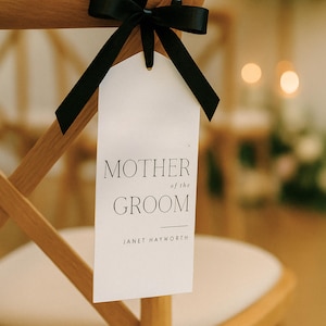 Op de afbeelding: Een houten stoel met een wit kussen en een label met de tekst "MOTHER of the GROOM" vastgebonden met een zwart lint. Het label bevat ook de naam "JANET HAYWORTH". De stoel staat in een goed verlichte kamer.