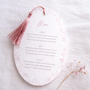 May include: A pale pink, oval-shaped menu card with a tassel. The menu features a floral border and a list of dishes, including starters, main courses, and desserts. The menu is printed in a delicate, elegant font.