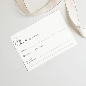 Puede incluir: Una tarjeta RSVP rectangular blanca con texto en negro que dice Kindly RSVP, By 1st January, Name(s), Accepts, Declines y Dietary Requirements, acompa&ntilde;ada de una cinta de color crema con un borde met&aacute;lico plateado fino en la parte superior.