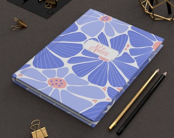 May include: A blue and white floral notebook with the word "Notes" on the cover. The notebook is lying on a brown leather surface with gold paper clips and pencils.