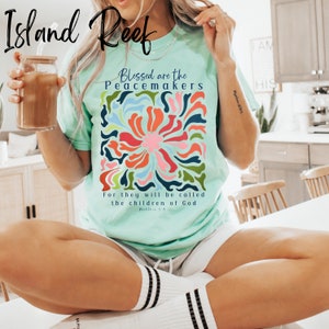May include: A light blue t-shirt with a colourful floral design and the text "Blessed are the Peacemakers" and "For they will be called the children of God" with a bible verse reference.