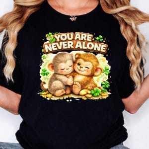 May include: Black t-shirt featuring a graphic of two cartoon monkeys hugging, surrounded by shamrocks and gold coins. The text "YOU ARE NEVER ALONE" is above the monkeys in a brown font.