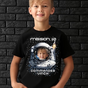 May include: Black t-shirt featuring a young boy in an astronaut helmet with the text "mission: 10" and "commander vinch". The helmet shows a view of Earth and a rocket ship blasting off. The design is in white and blue.