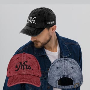 May include: Two distressed denim baseball caps with embroidered text. One cap is black with white text that reads "Mr. EST. 2025". The other cap is red with black text that reads "Mrs."