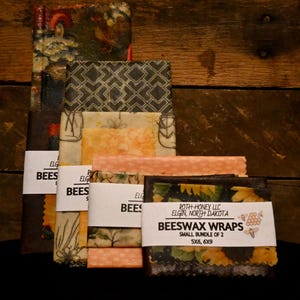 May include: A stack of colorful beeswax wraps with various patterns, including floral and geometric designs. The wraps are labeled with "BEESWAX WRAPS" and measurements of 5x6 and 6x9 inches. The background is a dark wooden surface.