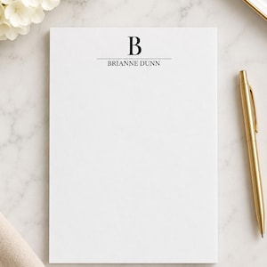 May include: A white rectangular notepad featuring a black serif letter B centred at the top above a horizontal line and the name BRIANNE DUNN in black text. A gold metal pen is positioned to the right of the notepad on a white surface.