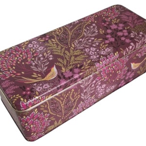 Puede incluir: Una caja rectangular de hojalata con un patrón floral en tonos de rosa, morado y dorado. La caja presenta un diseño de pájaro estilizado.