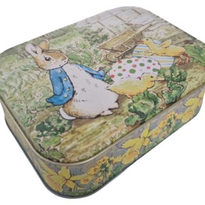 Puede incluir: Una caja de lata rectangular con una ilustración colorida de Peter Rabbit y pollitos. La caja tiene un fondo verde, marrón y amarillo con flores amarillas.