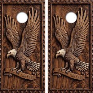 Könnte beinhalten: Zwei rechteckige Cornhole-Boards mit dunkelbrauner Holzmaserung. Jedes Board zeigt einen großen, detaillierten Adler mit ausgebreiteten Flügeln, der über einem Banner mit der Aufschrift LAND OF THE FREE thront. Ein Sternenrand umgibt die Kante.