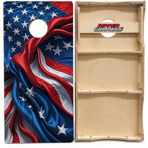 May include: A cornhole board featuring a printed graphic of a draped American flag with stars and stripes in red, white, and blue. The reverse side shows the natural light-colored wood construction with a Ripper Graphics logo printed on the support.