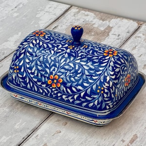 Op de afbeelding: Een blauwe keramische botervloot met een bloemmotief. Het deksel heeft een herhalend patroon van witte bladeren en oranje bloemen op een blauwe achtergrond. De schaal staat op een zilverkleurige metalen lade.