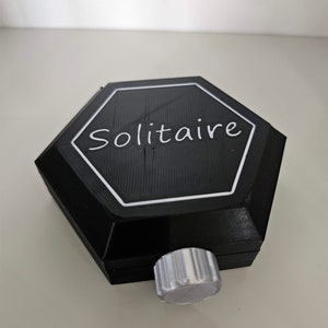 May include: A black hexagonal game box with a white inscription that reads "Solitaire". The box has a silver knob on the bottom.