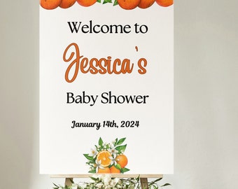 May include: White welcome sign with orange blossoms and oranges at the top and bottom. The sign reads "Welcome to Jessica's Baby Shower January 14th, 2024".