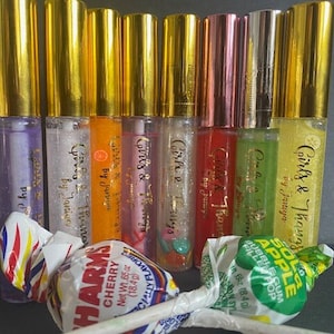 Peut inclure: Huit tubes de gloss à lèvres transparents avec des bouchons et des étiquettes dorés. Les étiquettes portent une inscription dorée "Girls & Thangs" et "by Janiya". Les tubes sont disposés en rangée sur une surface.