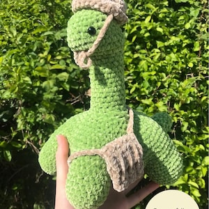 May include: Green crocheted dinosaur wearing a brown crocheted hat and a brown crocheted vest. The dinosaur is being held by a hand. The text "Easy to follow PDF pattern with photo guides!" is in the bottom right corner of the image.