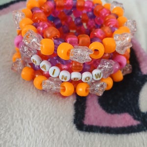 Puede incluir: Una pulsera de cuentas multicolores con la palabra "RAINBOW" escrita en cuentas blancas y doradas. La pulsera presenta cuentas naranjas, rosas, moradas y transparentes. Un accesorio vibrante.