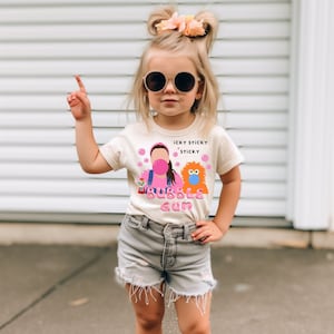 May include: A young girl wearing a white t-shirt with a pink and orange cartoon character design. The shirt says "icky sticky sticky bubble gum". She is wearing denim shorts and sunglasses.