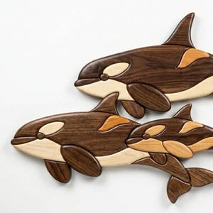 May include: Three wooden orca whale wall art pieces. The whales are crafted from various wood tones, including dark brown, light brown, and tan. The orcas are arranged in a family group, with a large whale, a medium whale, and a small whale.